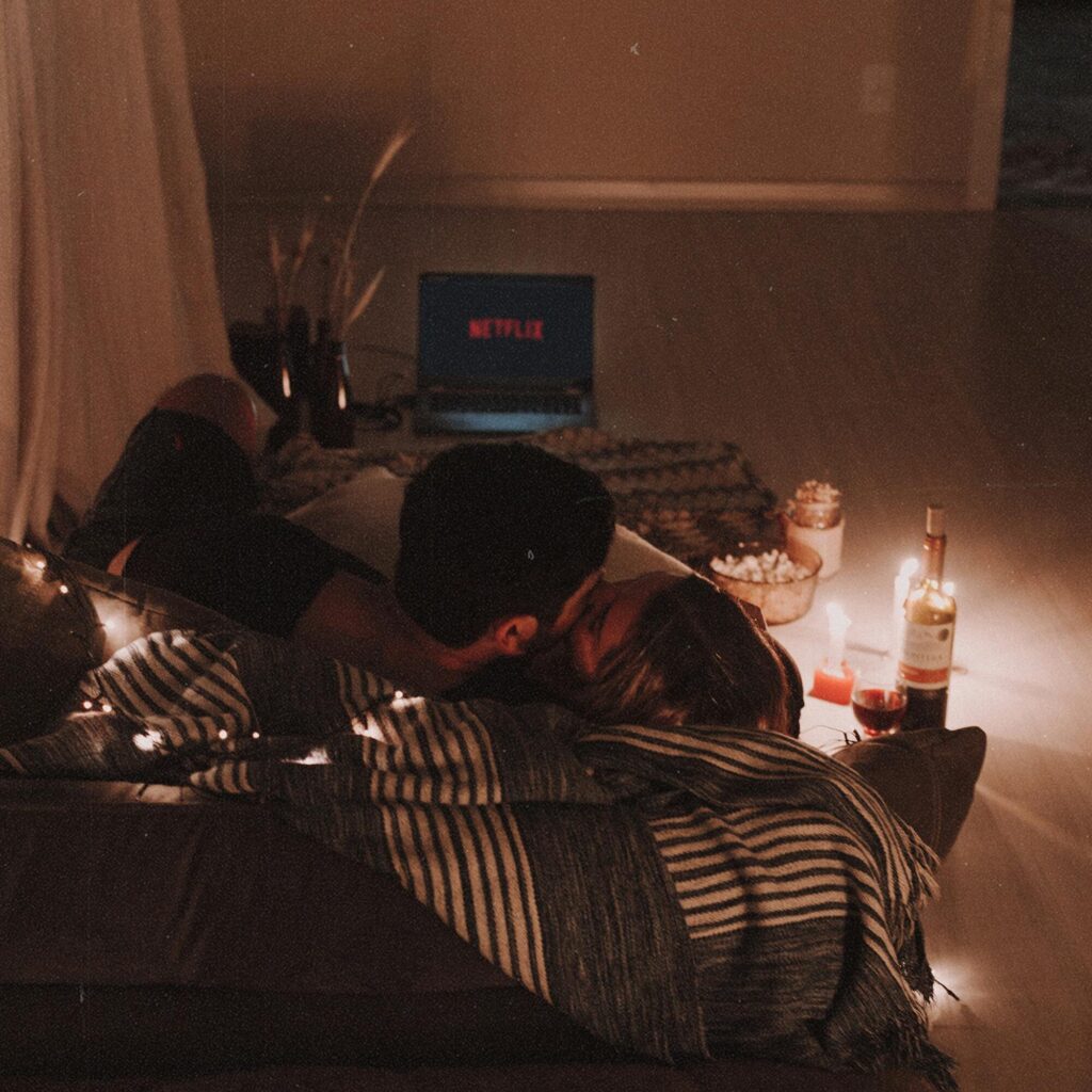 Cozy indoor movie night setup with a couple cuddling on a couch, surrounded by candles and wine.